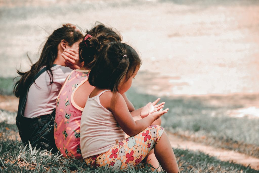 three little girls sitting in a row on a hill one with her hands out as she talks